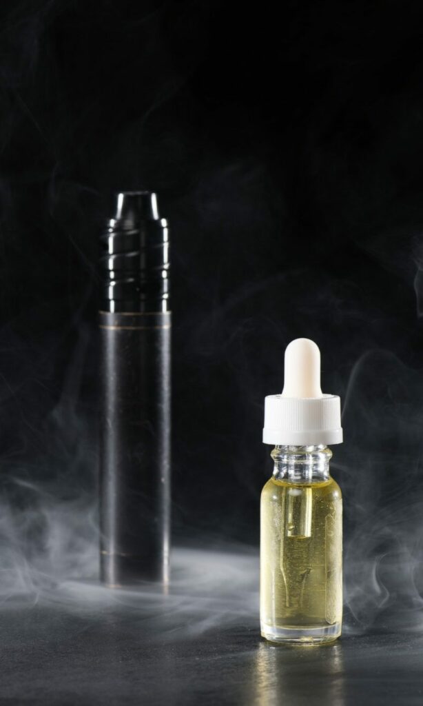 A bottle of vaping liquid with a dropper cap in front of a dark-colored vape device, both surrounded by smoke against a dark background.