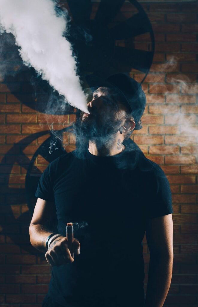 A person wearing a black hat and shirt exhales a large cloud of vapor while holding a vaping device against a brick wall backdrop.