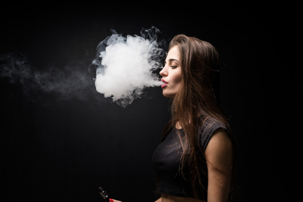 A woman with long hair wearing a black crop top exhales smoke while holding a vaping device against a black background. Explore Affordable vape products at Vape World GA.