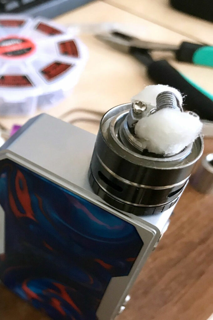 Close-up of a vape device with the coil and cotton wick exposed. Tools and a spool of wire are seen in the background, showcasing genuine vape devices from an Athens GA vape store offering low-cost vape options.