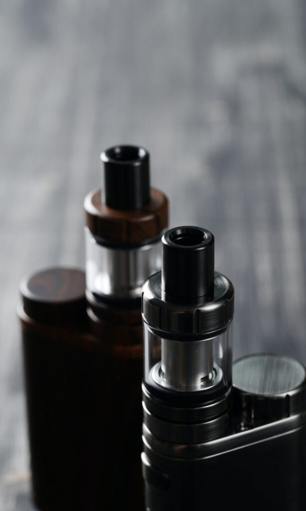 Close-up of two electronic vaping devices with black mouthpieces on a dark, blurred background. One device has a wooden finish, while the other has a metallic finish.