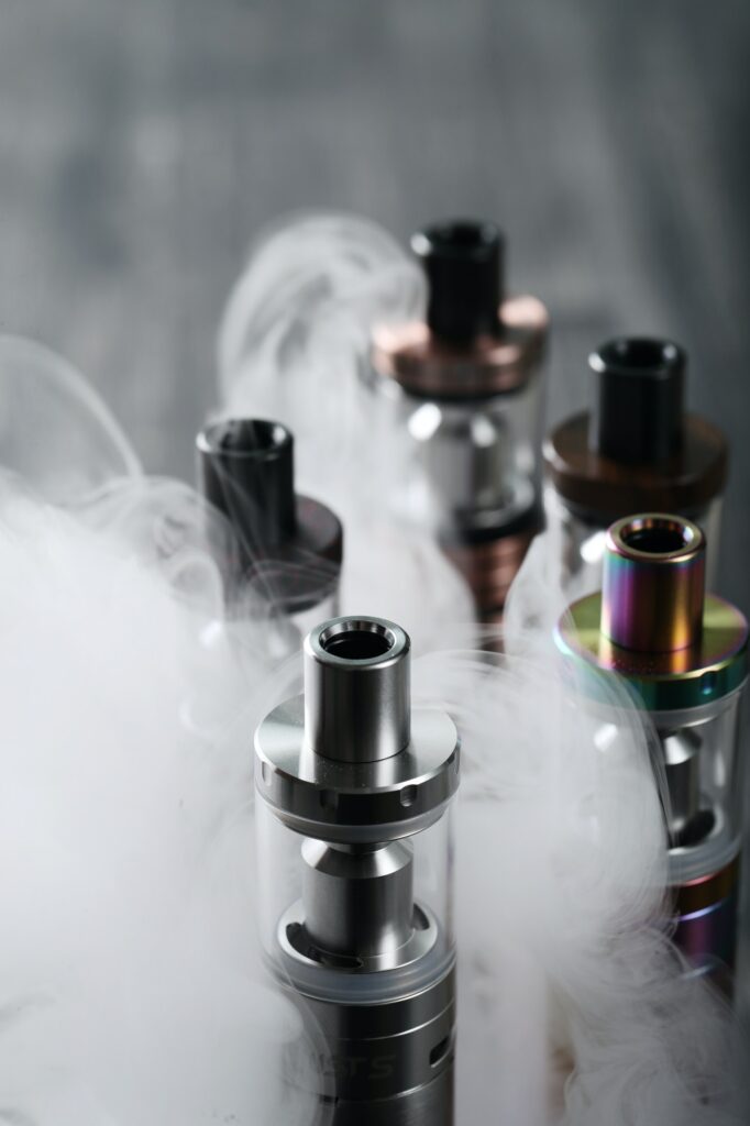 Several vaping devices with metallic finishes emit vapor streams, set against a blurred background.