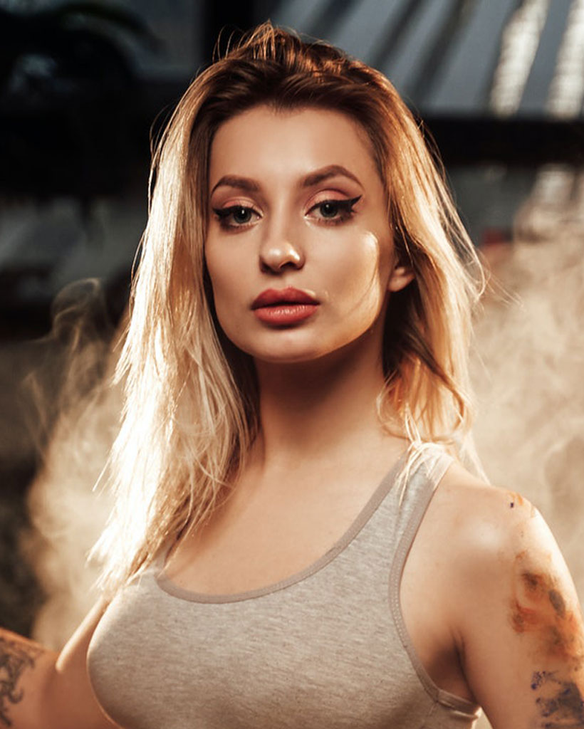 A woman with long blonde hair and a serious expression stands in a smoky background, wearing a grey tank top, representing the quality of Genuine vape devices from Vape World GA.