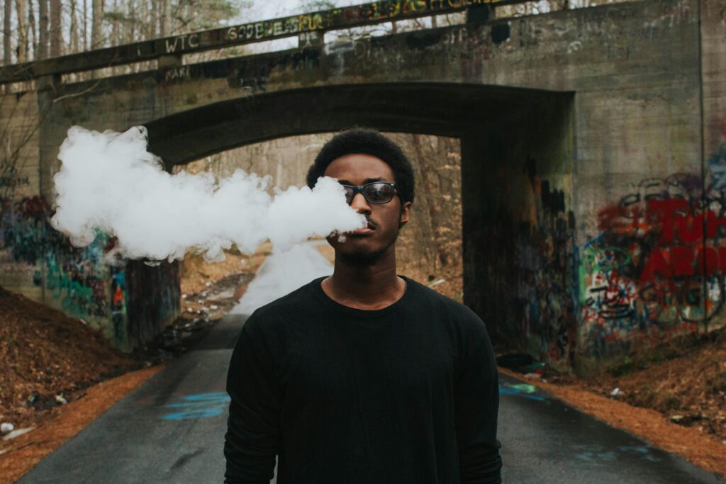 A person wearing glasses blows out a large cloud of vapor while standing on a graffiti-covered bridge, having just visited a local vape store.